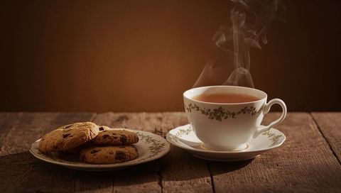 Steaming china teacup with floral saucer and chocolate chip cookies on rustic wood cozy still life