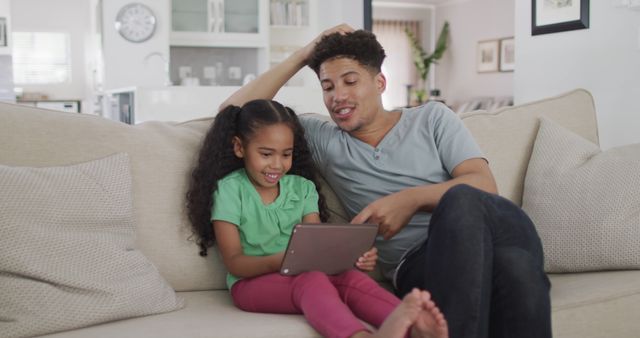 Father and daughter enjoying time together on couch, engaging with tablet. Used for themes of family bonding, technology in daily life, and modern parenting. Ideal for educational, technological, and family-oriented content.