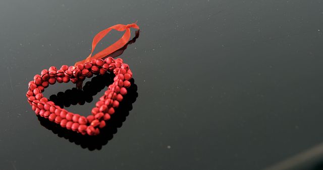A red beaded heart with a ribbon lies on a reflective black surface, with copy space. Its vibrant color and heart shape symbolize love and affection, making it suitable for themes like Valentine's Day or romantic celebrations.