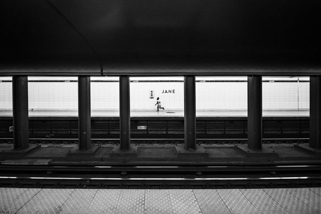 A lone commuter is walking across a subway platform, with the name 'Jane' on the wall in a minimalistic, black-and-white urban setting. This image captures the solitude and architectural elements of urban transport systems. It can be used for topics related to travel, urban lifestyle, public transportation, minimalism, and modern cityscapes.