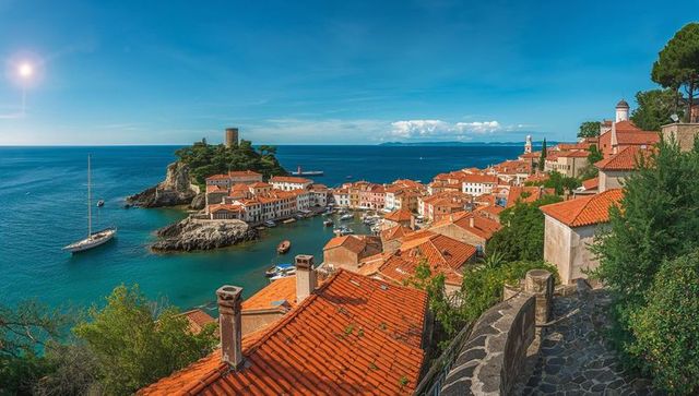 Sunlit mediterranean harbor with terracotta roofs cascading to turquoise bay and sailboats