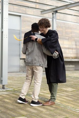Diverse friends celebrating graduation hug on campus courtyard with gown and mortarboard