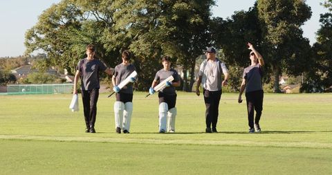 Diverse male cricketers walking onto pitch carrying bats and pads with coach leading team