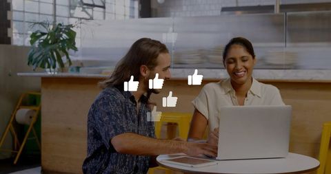 Coworking duo creating social media thumbs-up graphics on laptop in casual cafe teamwork