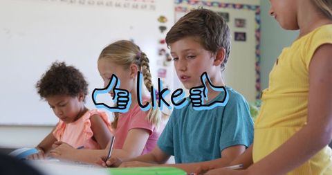 Elementary students writing at classroom table with like thumbs-up overlay suggesting engagement