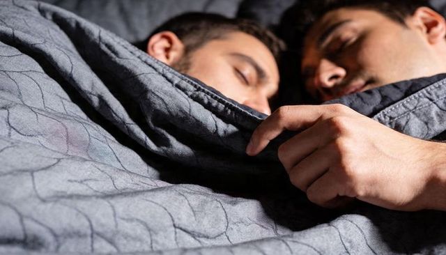 Two men sleeping close under gray quilted blanket holding edge intimate bedroom moment