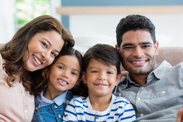 Happy family sitting comfortably on a sofa, smiling and looking at camera. Ideal for use in family-oriented advertisements, articles about parenting, home lifestyle content, and brochures promoting family services or products.