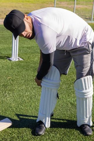 Man in 30s fastening cricket batting pads on artificial turf during outdoor practice