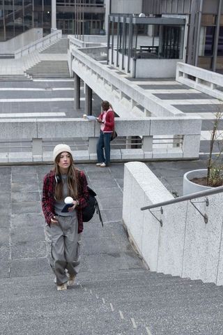 Young woman climbing concrete plaza stairs carrying coffee and backpack, urban commuter
