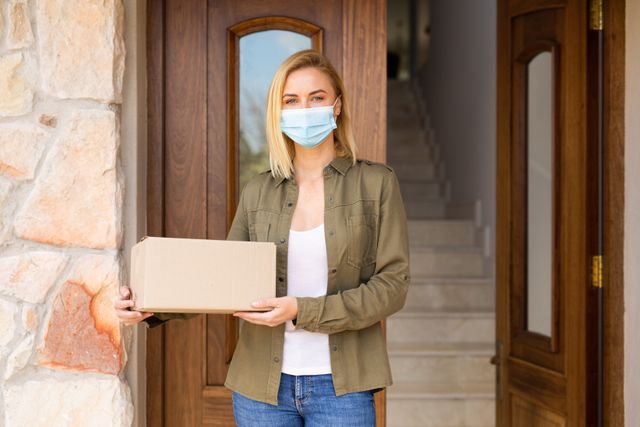 Caucasian woman wearing a mask standing outside the front door of her house while holding a package. behind her is an open door leading to the stairs.
