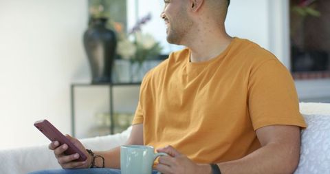 African man relaxing on sofa with smartphone and coffee mug wearing mustard t-shirt