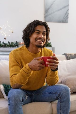 Young indian man sitting on sofa holding red mug smiling in cozy living room