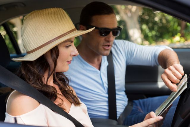 Couple sitting in car, reading map, planning route for road trip. Man pointing at map, woman wearing hat and seatbelt. Ideal for travel blogs, vacation planning, road trip guides, transportation services, and adventure-themed promotions.