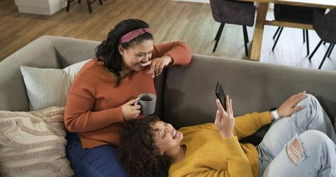 Mother and daughter lounging on couch sharing smartphone and coffee, laughing together