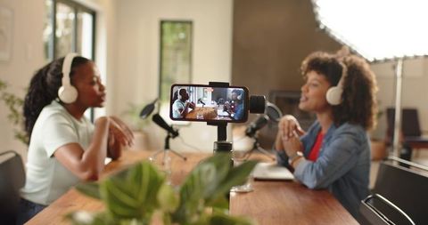 Diverse women recording podcast with headphones, smartphone gimbal, microphones, home studio