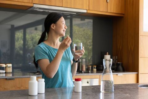 Asian woman taking daily medication in modern kitchen morning routine with water glass