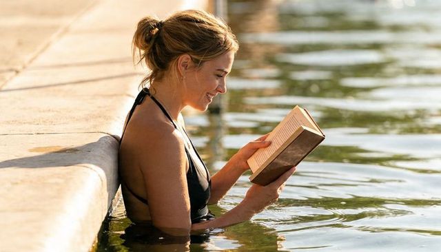 Sunlit woman reading paperback while relaxing in pool by concrete ledge, summer leisure