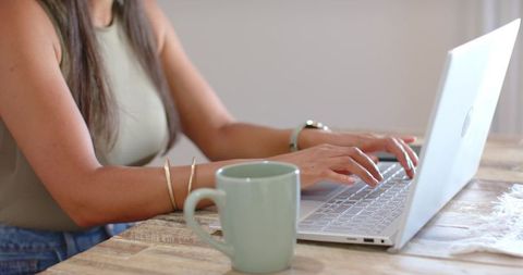 African american woman typing on laptop at wooden table with mint mug and gold bracelets