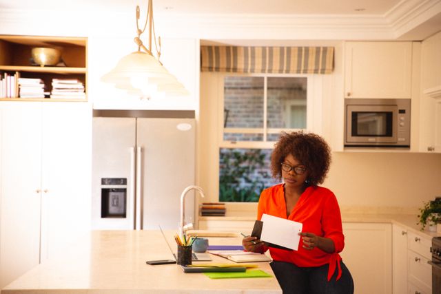 Happy african american woman sitting, working in kitchen. technology and communication, flexible working from home.