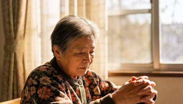 Senior asian woman contemplating by sunlit window at home wearing floral cardigan