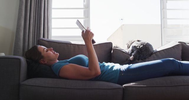 Woman lying back on gray couch reading a tablet while a black dog rests beside her. Sunlight streams in through window creating a peaceful, comfortable scene. Perfect for themes of relaxation, leisure activities, home comfort, and companionship with pets.