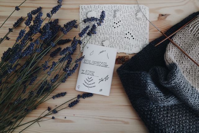 Handmade coloring zine, dry lavender flowers, drawing, and knitwear on wooden table. Ideal for showcasing crafts, homemade products, creative projects, and artistic activities in a rustic setting.