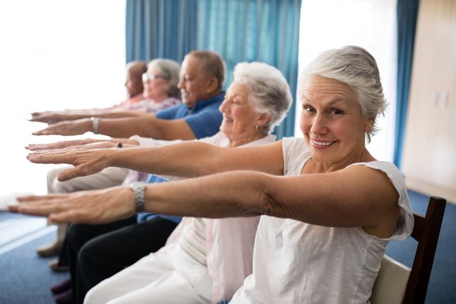 Senior individuals are seen exercising together in a retirement home. They are seated and stretching their arms forward, smiling and enjoying the activity. This image can be used for promoting senior wellness programs, community activities, and active lifestyles for the elderly.