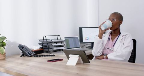 Clinician reviewing charts while sipping coffee at modern clinic desk with tablet and laptop