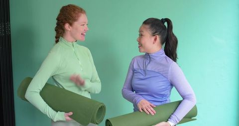 Caucasian and asian women holding yoga mats chatting and smiling in mint fitness studio