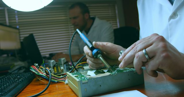 Close-up of technicians working on circuit board repair with a soldering iron at a workstation. One technician using the soldering iron to fix electronics, while the other is in the background, focused on their task. Ideal for technology and engineering concepts, illustrating precision work, and showcasing expertise in electronics repair.