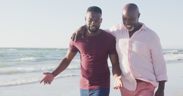 Two men, likely father and son, are walking along the beach, arm in arm, enjoying a sunny day by the ocean. They are engaging in conversation and smiling, demonstrating a strong and happy family bond. This image can be used for themes such as family relationships, happiness, summer vacations, and spending quality time outdoors.