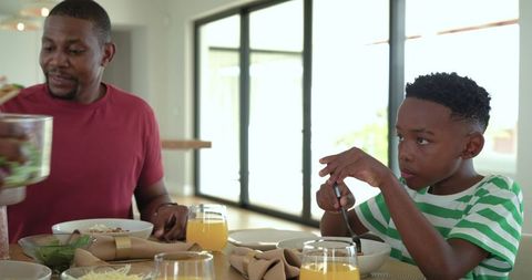 African american father and son sharing sunlit breakfast at home dining table with orange juice