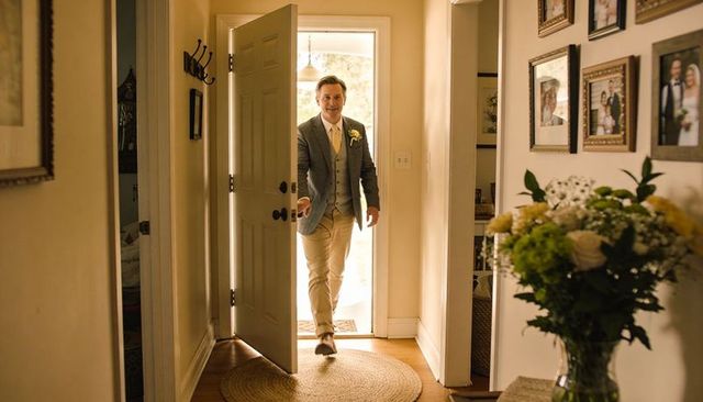 Man entering sunlit home foyer wearing suit and boutonniere, warm welcome and floral accents