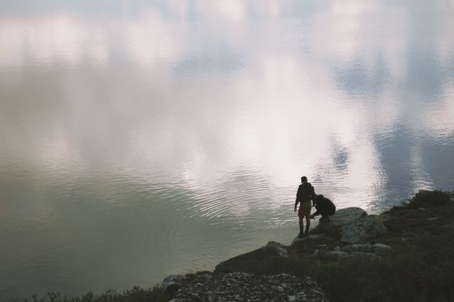 Man stands beside peaceful lake at dawn, clouds reflecting on water, perfect for themes of tranquility, solitude, connection with nature, adventure lifestyle, travel ads and outdoor magazines.