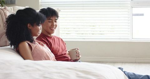 Cozy mixed-race couple relaxing beside bed with morning coffee in minimalist bedroom
