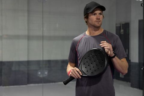 Focused padel player standing on indoor court holding perforated paddle wearing cap
