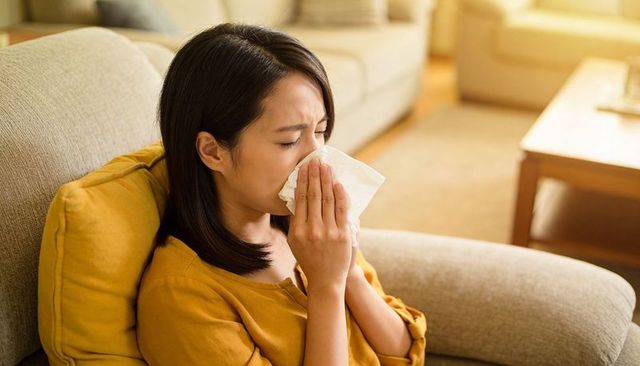 Asian woman sneezing on sofa at home holding tissue, allergy cold, resting on couch