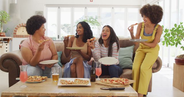 Happy diverse female friends sitting on sofa in living room, eating pizza. spending quality time at home.