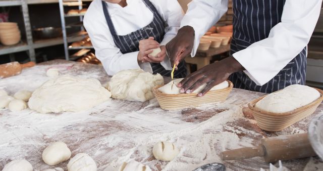 This stock photo features two professional bakers working together in an artisan bakery kitchen, focusing on shaping bread dough. One baker is cutting patterns into the dough while the other kneads it. Perfect for marketing materials related to baking, culinary arts, teamwork in the kitchen, and artisanal food production.