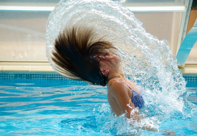 This vibrant image depicts a woman flipping her hair back in an outdoor swimming pool, creating a beautiful water splash. The scene captures joy and summertime fun, making it ideal for use in advertisements focusing on leisure, vacation, or health and wellness. It can also be utilized in editorials or social media posts promoting swimming pools, swimwear, or outdoor activities.