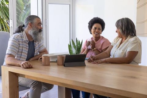 Three adults chatting around wooden table with tablet and mugs, casual home meeting