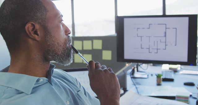 An architect sits in his office thoughtfully planning a building blueprint displayed on his computer screen. His workspace features technical drawings and design notes. Perfect for business websites, architecture firms, and engineering publications.