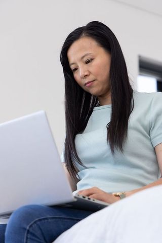 Asian woman working on laptop in bed wearing wristwatch and light blue top