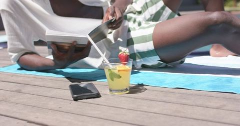 Man reading paperback on pool deck towel with refreshing citrus cocktail and smartphone