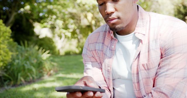 Young man wearing a plaid shirt and white t-shirt is looking at his smartphone while sitting in a lush garden. Trees and shrubbery provide a serene backdrop. This image is ideal for use in ads or blogs about outdoor activities, modern technology, casual fashion, or lifestyle content.