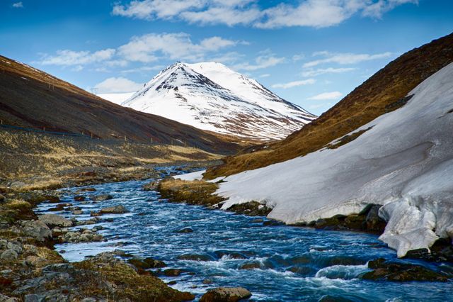 Majestic mountain landscape featuring a snow-capped peak, flowing river, and blue sky. Ideal for travel blogs, adventure promotions, nature calendars, and outdoor activity advertisements.