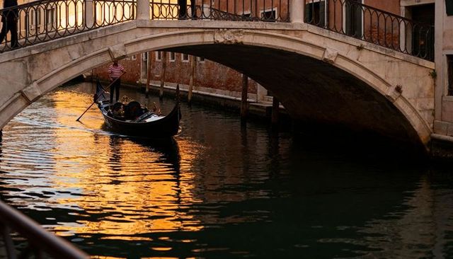 Gondolier steering gondola under arched bridge on venice canal at sunset with golden reflections