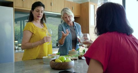 Women chatting over wine at sunlit kitchen island sharing salads and fruit bowl