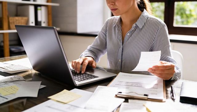 Working from home organizing receipts and invoices on laptop and clipboard at bright desk
