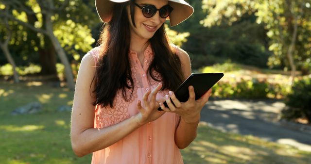 Young woman standing in park using tablet on a sunny day, wearing wide-brimmed hat and sunglasses. Suitable for articles about outdoor activities, technology, digital communication, new age lifestyle, and fashion.